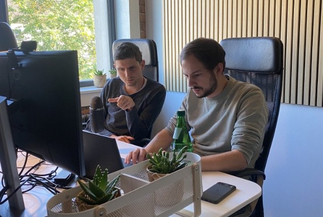 Thomas und Christopher am Schreibtisch sitzend, diskutierend und auf Bildschirme blickend.