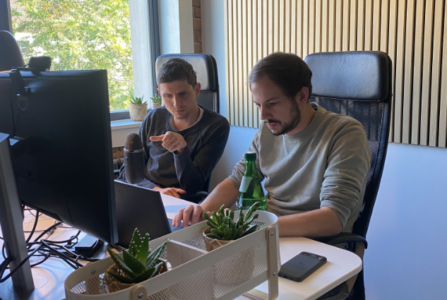 Thomas und Christopher am Schreibtisch sitzend, diskutierend und auf Bildschirme blickend.