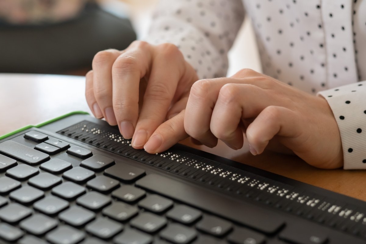Eine blinde Frau benutzt einen Computer mit Braillezeile und Computertastatur. Inklusive Gerät.